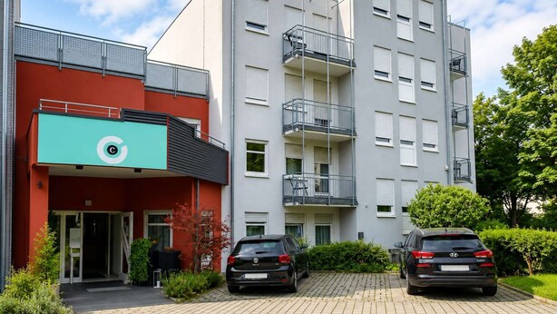 En flereetagers boligbygning med rød facade, en grøn markise med et logo, og to biler parkeret foran.
