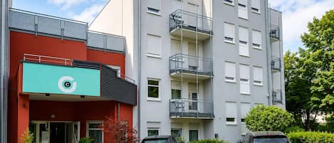 A multi-story residential building with a red facade, a green awning with a logo, and two cars parked in front.