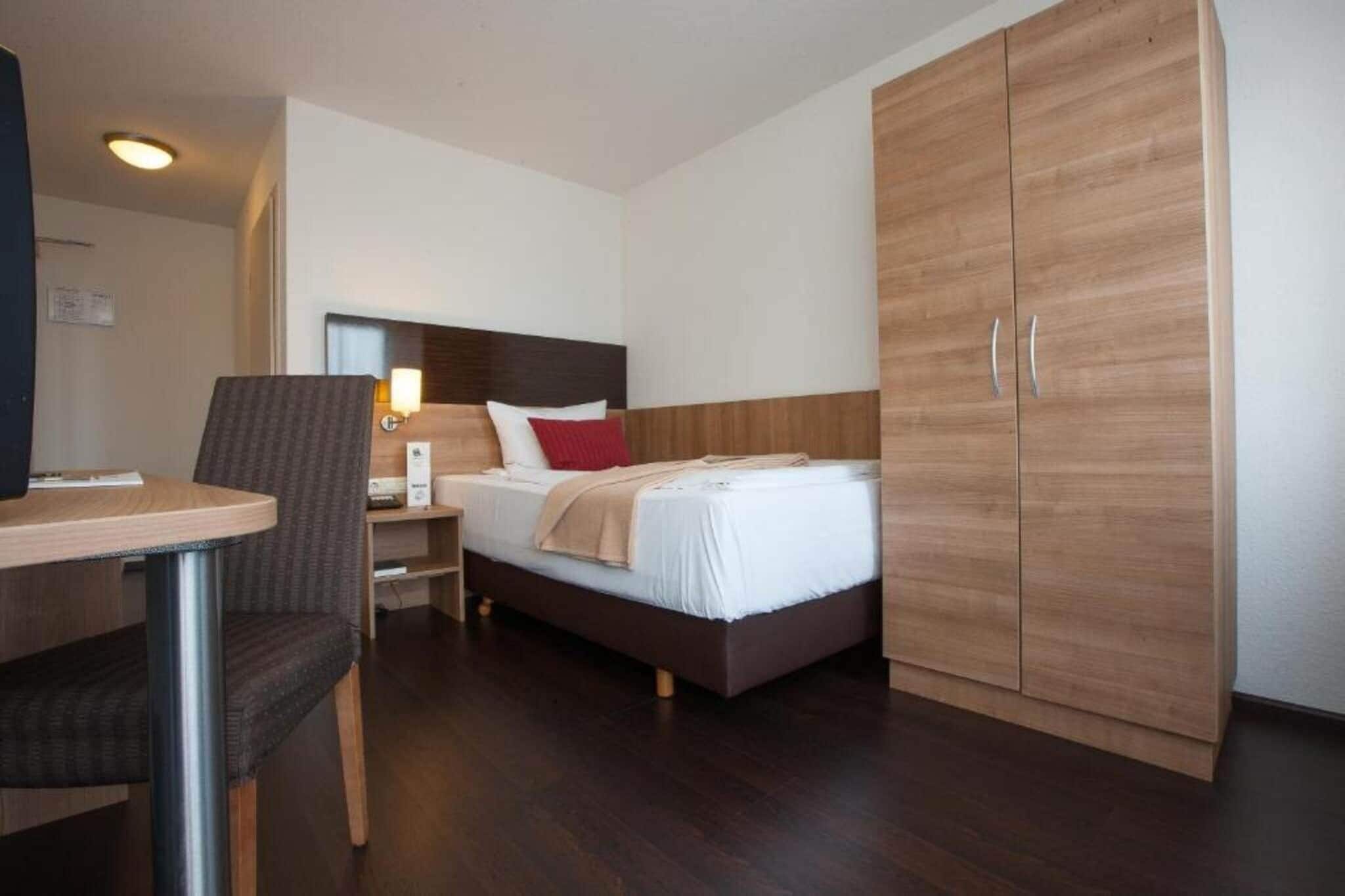 Una habitación de hotel con cama, escritorio, silla, armario y mesita de noche.