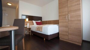Una habitación de hotel con cama, escritorio, silla, armario y mesita de noche.