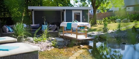 Un entorno tranquilo al aire libre con un pequeño estanque, una terraza de madera con una mesa y un edificio moderno con una zona cubierta.