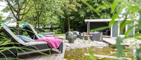 Una terraza con sillones reclinables y un pequeño estanque, rodeada de árboles y un espacio de descanso techado.