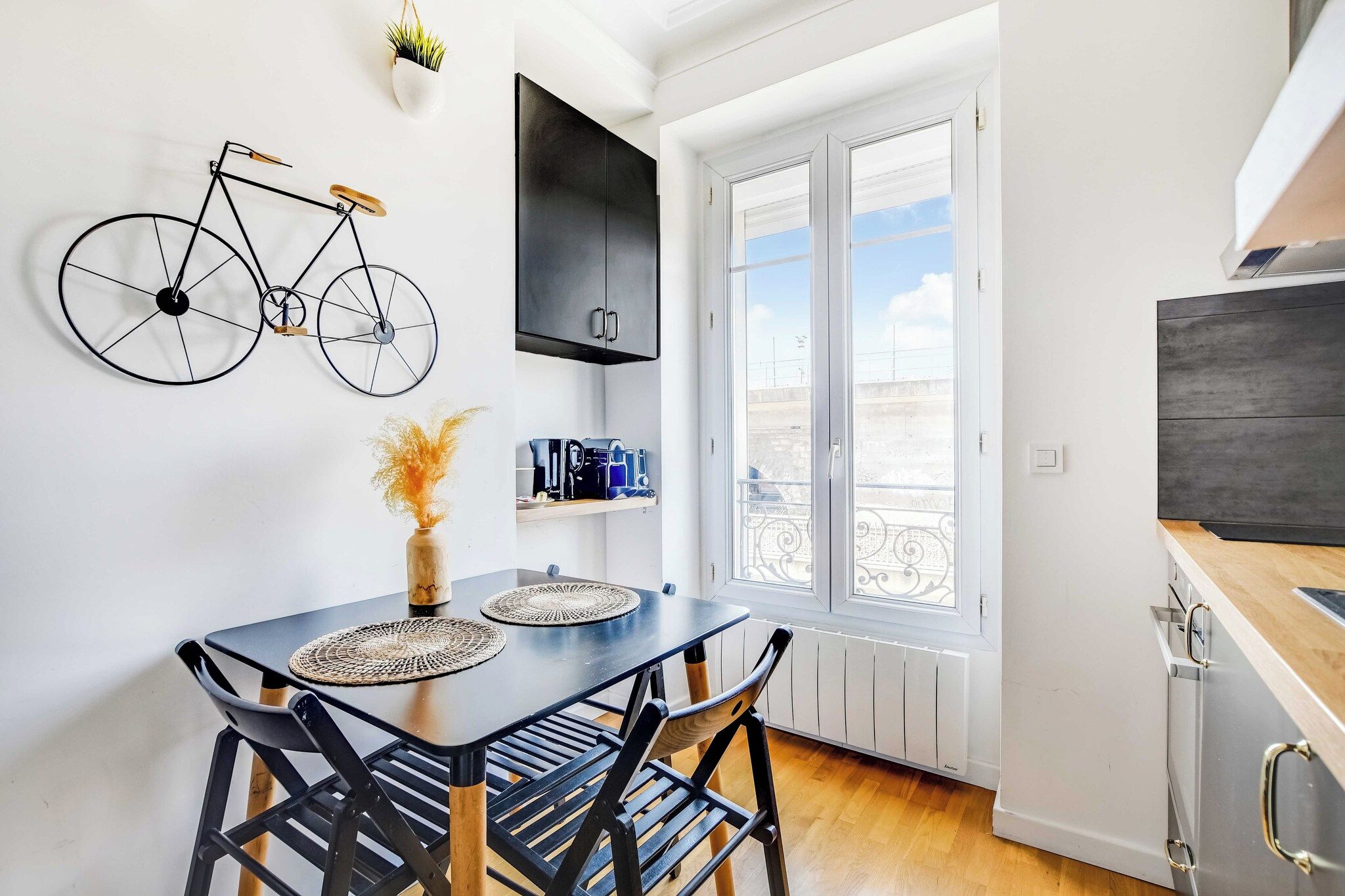 Eine moderne Küche mit Essbereich, ausgestattet mit einem schwarzen Esstisch und gewebten Tischsets, einem an der Wand befestigten schwarzen Fahrrad und Blick auf die Stadt durch große Fenster.