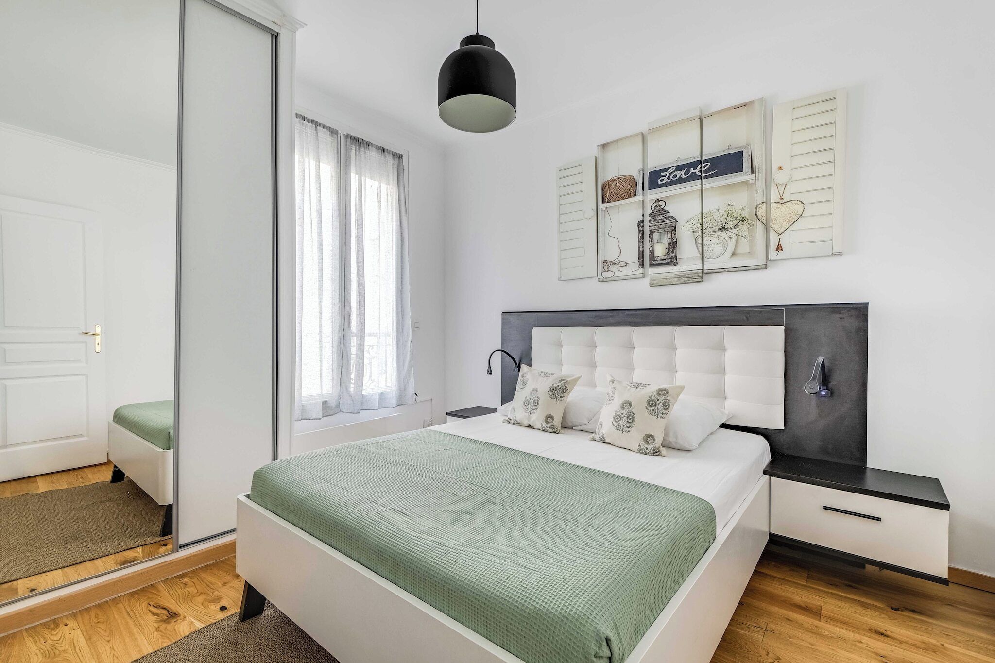 A bedroom with a bed, bedside tables, a hanging light fixture, a window with curtains, and wall decorations.
