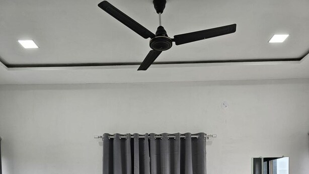 A bedroom with a bed, a ceiling fan, a window, and a nightstand. - Hotel Randha (Akola)