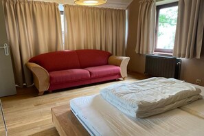 Une pièce avec un canapé rouge, un lit avec un matelas blanc, un plancher en bois et des rideaux de couleur beige.