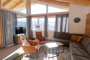 Un salón acogedor con un sofá, sillones, una mesa de centro y un televisor. Ventanas amplias permiten la entrada de luz natural y vistas al exterior.