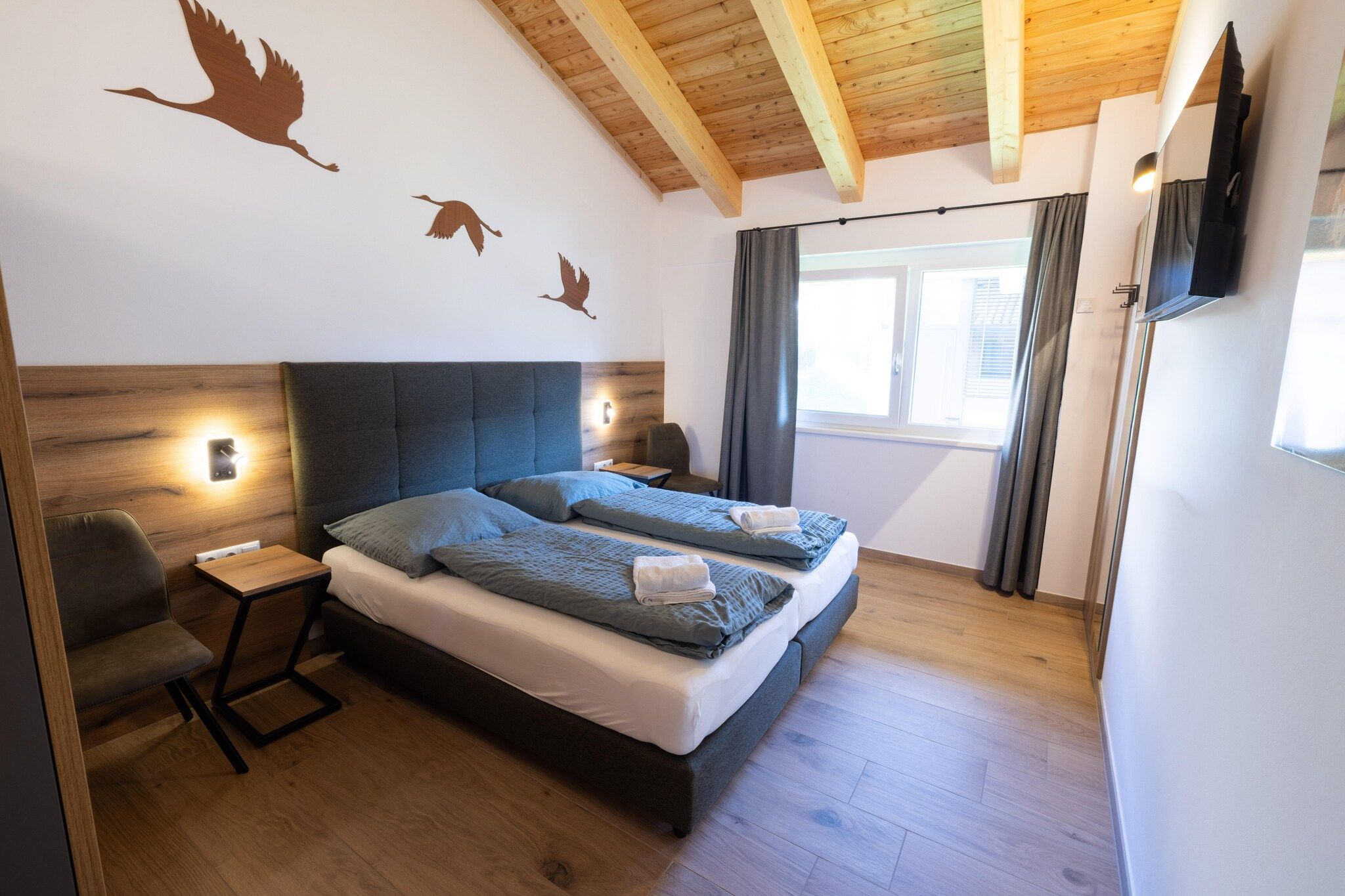 A modern bedroom with a large bed, a desk, and a chair. There are wall-mounted lights and a TV.