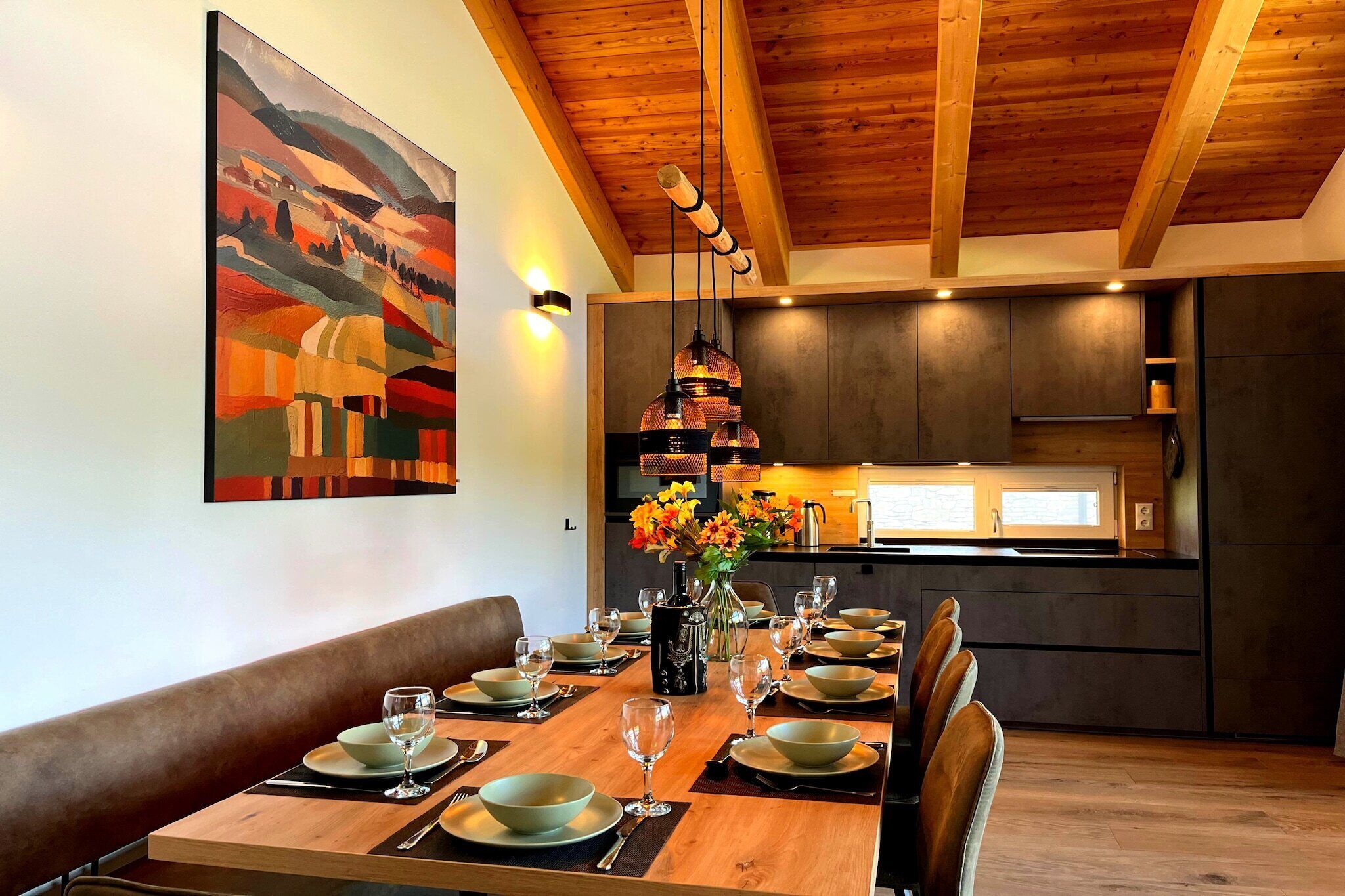 Un coin repas avec une table en bois dressée pour six personnes, un vase rempli de fleurs et un tableau accroché au mur.