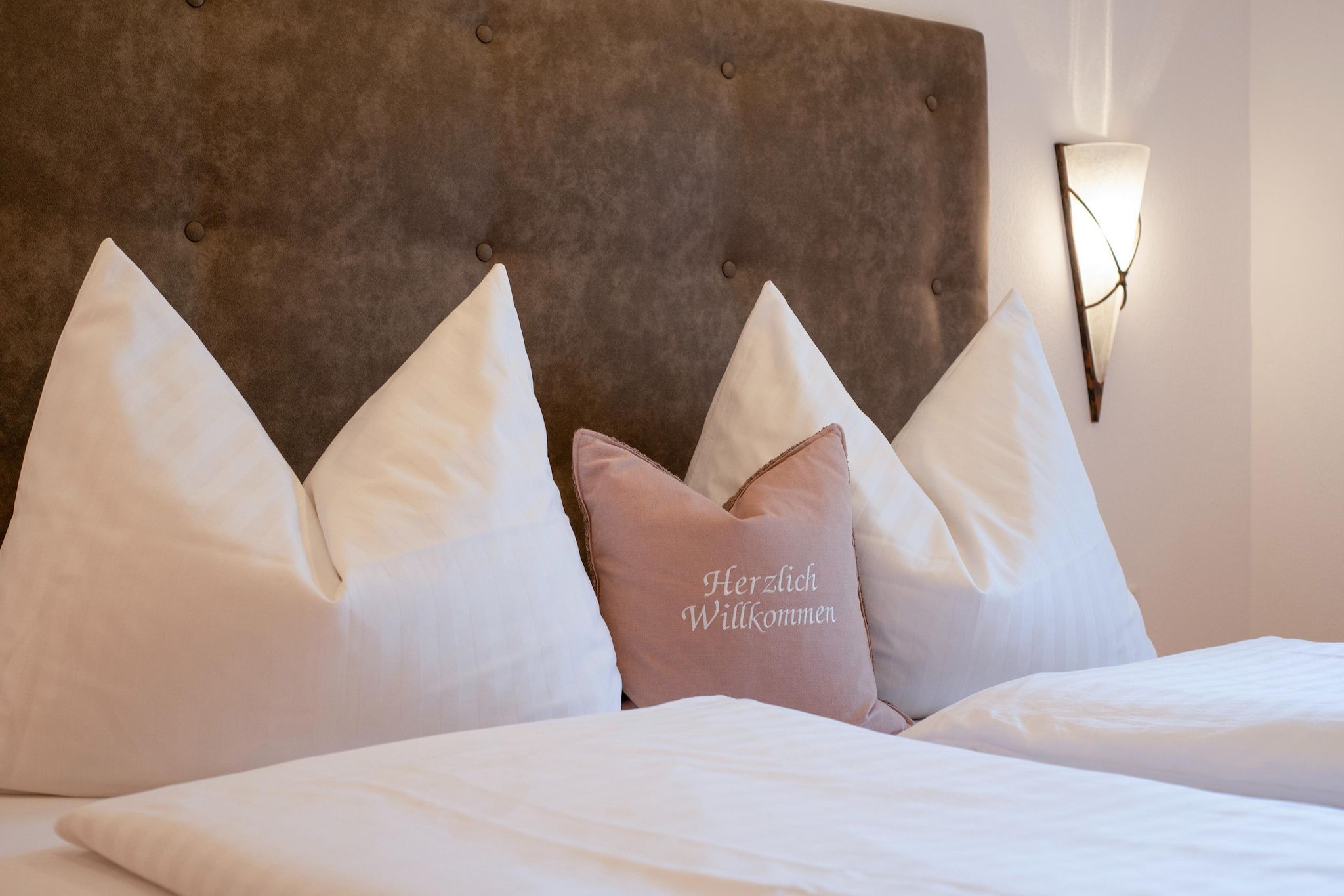 Cama com roupa de cama branca e cabeceira marrom. Há um luminária fixada na parede.