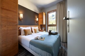 Ein Hotelzimmer mit Bett, Holz-Kopfteil, zwei Wandlampen, einem Fenster mit Vorhängen und Blick ins Freie.