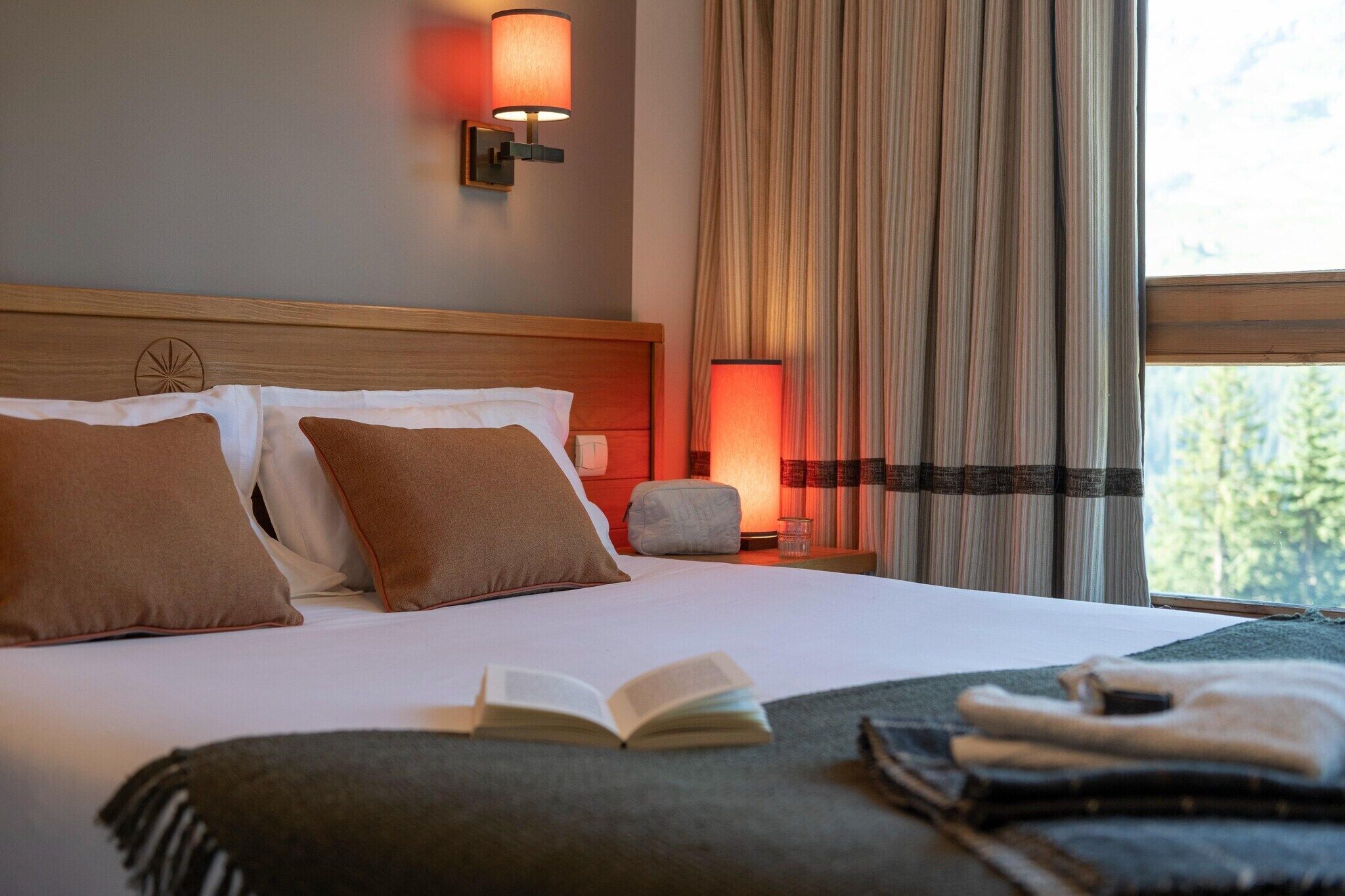 Habitación de hotel con cama, almohadas, mesita de noche con lámpara, ventana con cortinas y vista a árboles.