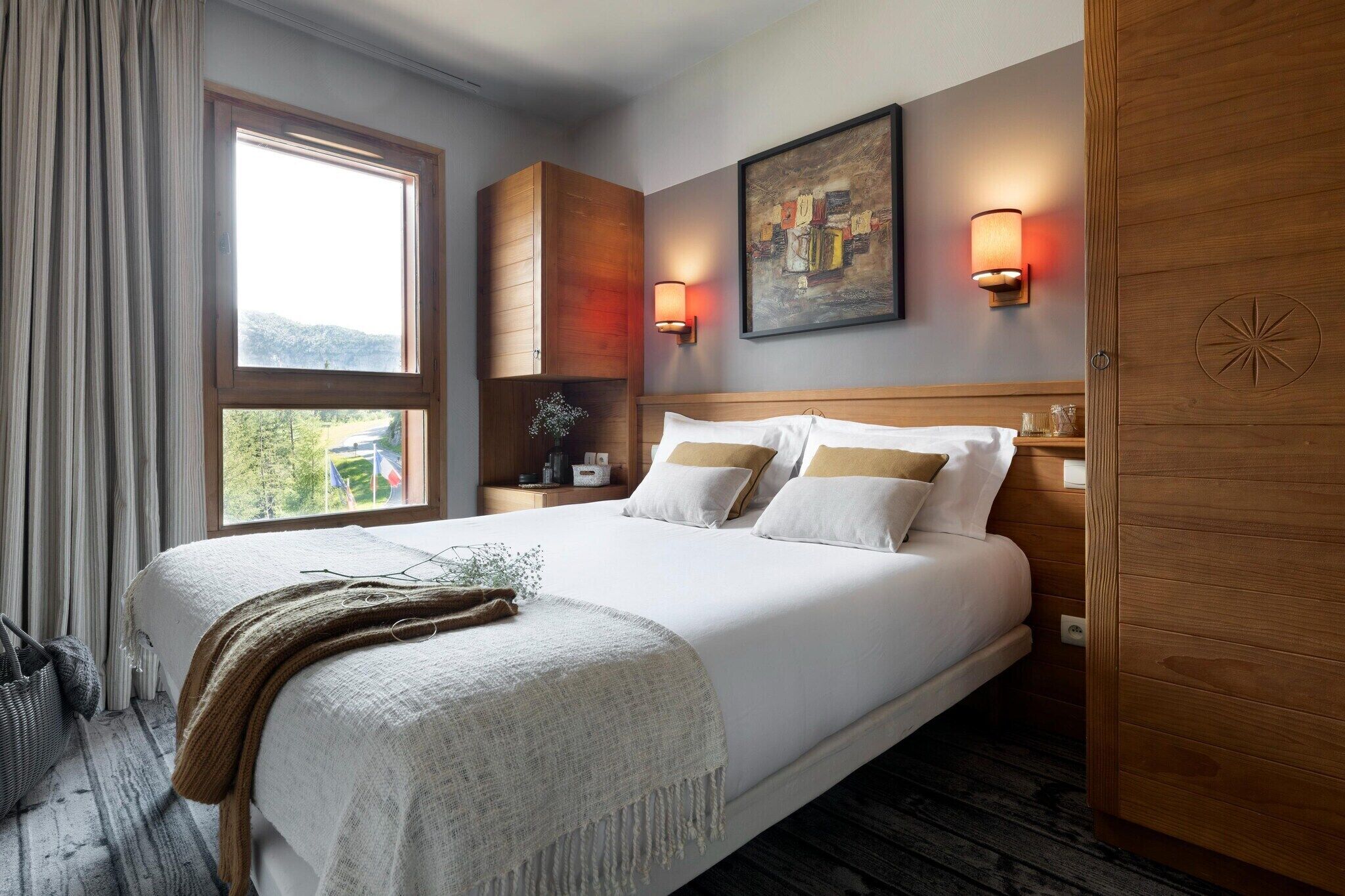 Una camera d'albergo con un letto grande, un comodino, una finestra con tende, un quadro appeso al muro e una porta in legno.