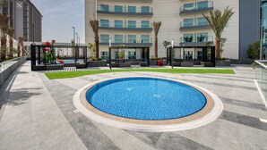 Una piscina con borde de azulejos, rodeada de un área pavimentada con palmeras y un edificio al fondo.