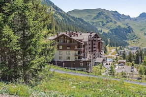 A multi-story building with balconies, surrounded by lush greenery and mountains. - Luxury 10-Person Alpine Panoramic Suite (Flaine)