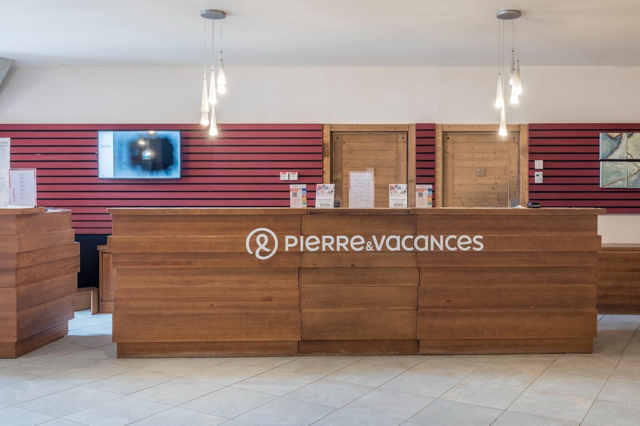 A reception desk with a logo, a mounted TV, and hanging pendant lights.