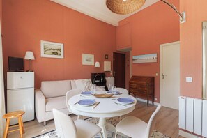 A room with a round dining table set for four, a sofa, a television, and a radiator. - Flat in a traditional Norman townhouse – Garden (Deauville)