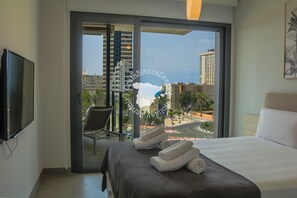 Quarto de hotel com cama, televisão, poltrona e vista da cidade por uma porta de vidro.