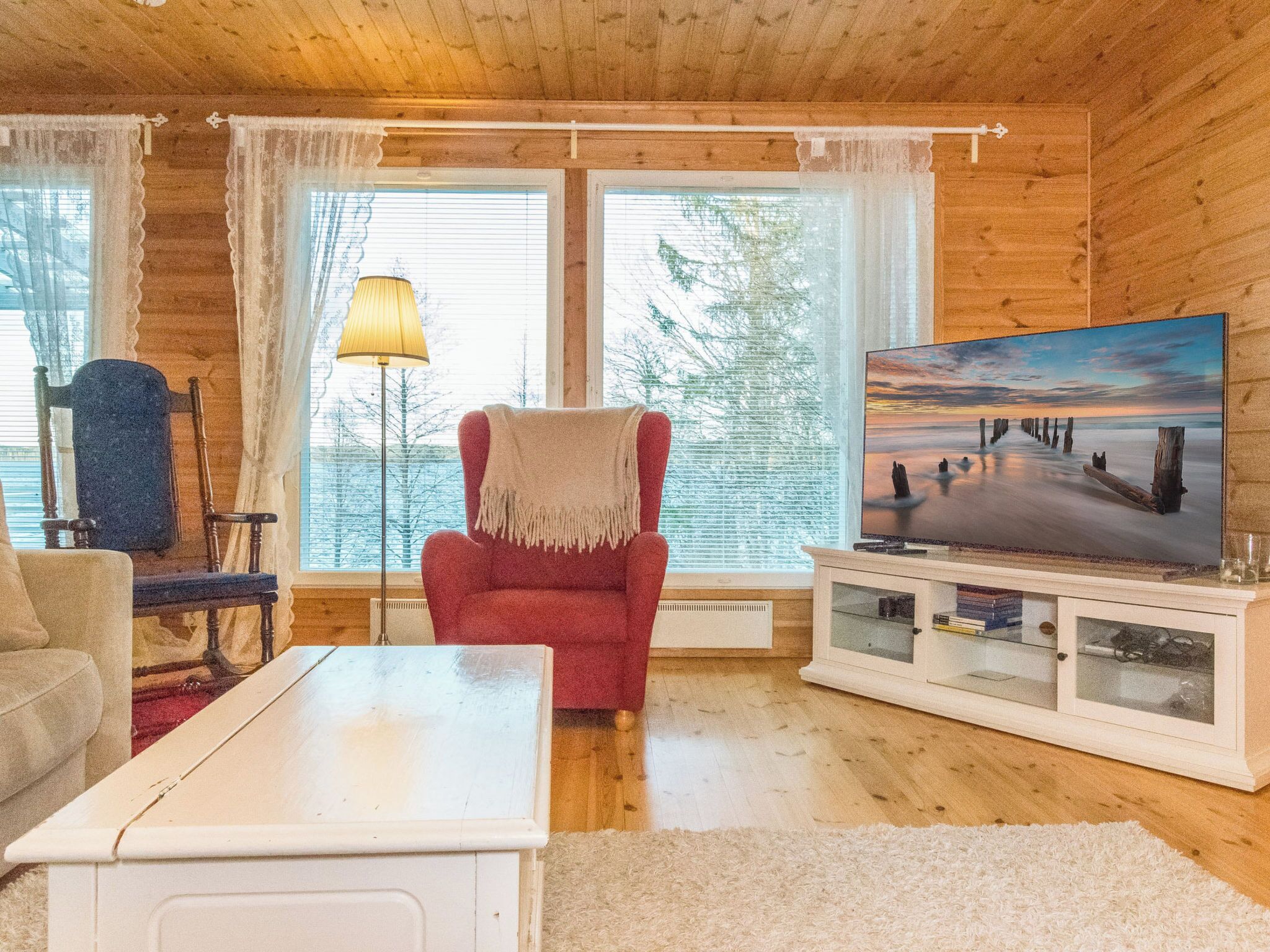 A cozy cabin interior with a red armchair, a white coffee table, a floor lamp, and a television on a cabinet.