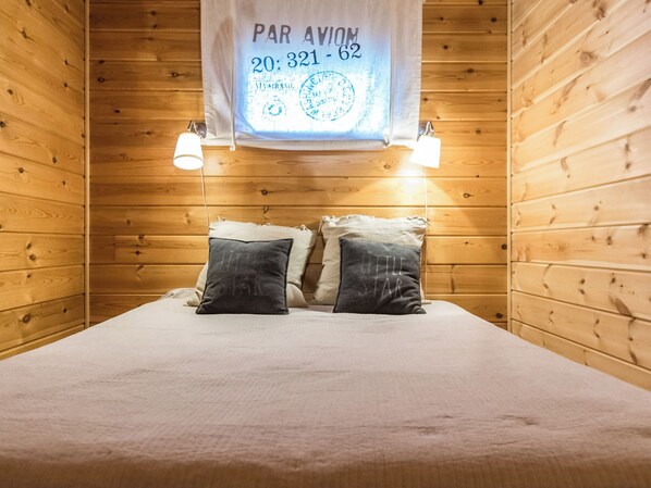 A wooden room with a bed, two pillows, a hanging sign with text, and two wall-mounted lights.