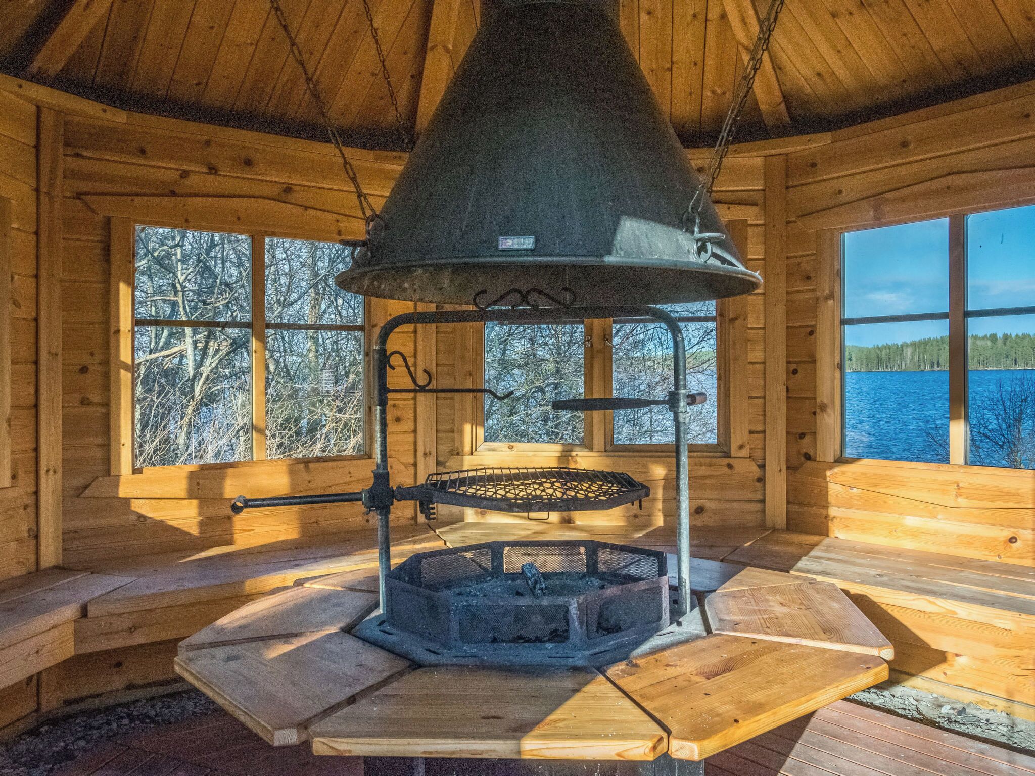 A wooden cabin with a central fireplace, large windows, and a view of a lake.
