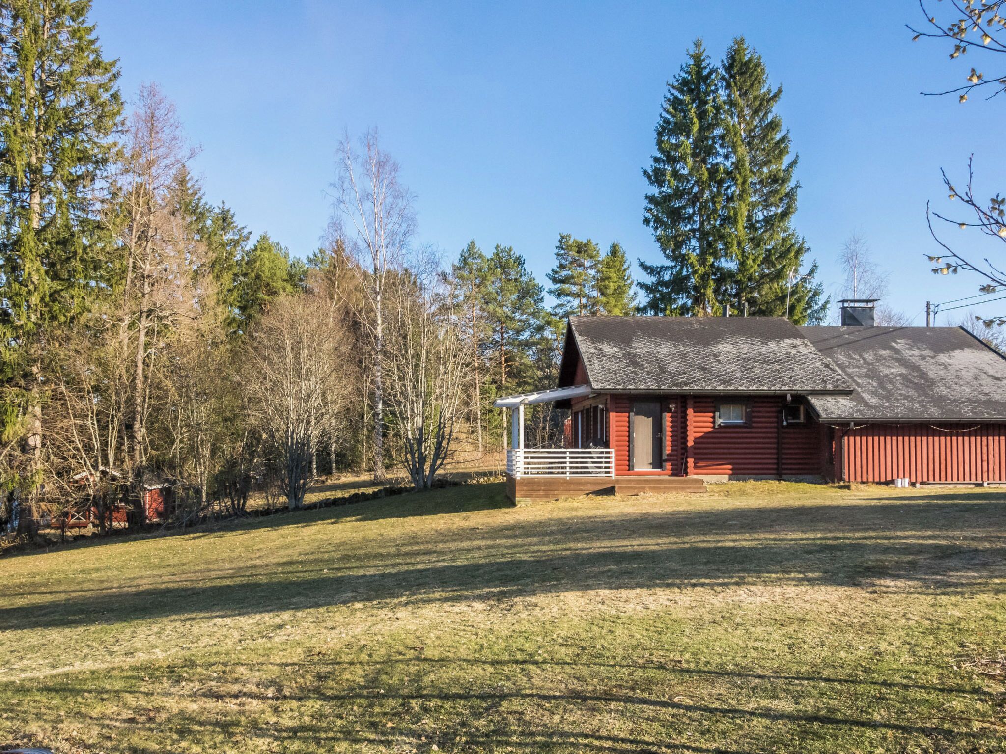 A red wooden cabin with a sloped roof, surrounded by a grassy field and tall trees.