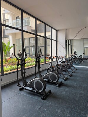 A gym with stationary bikes and treadmills, large windows, and a view of a building outside. - Modern Luxury Apartment | Sauna, Pool, Gym & Balcony (Nairobi)