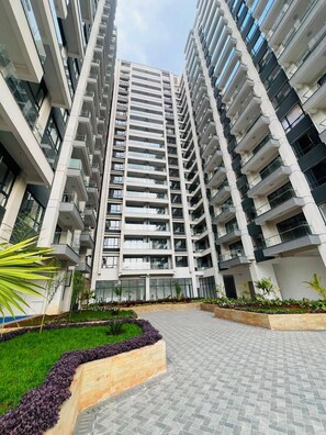 Un immeuble résidentiel de grande hauteur doté de nombreux balcons, d’une promenade dallée et d’espaces paysagers avec des plantes.