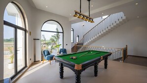 Een ruime kamer met een pooltafel, een trap en grote ramen met uitzicht op de omgeving.