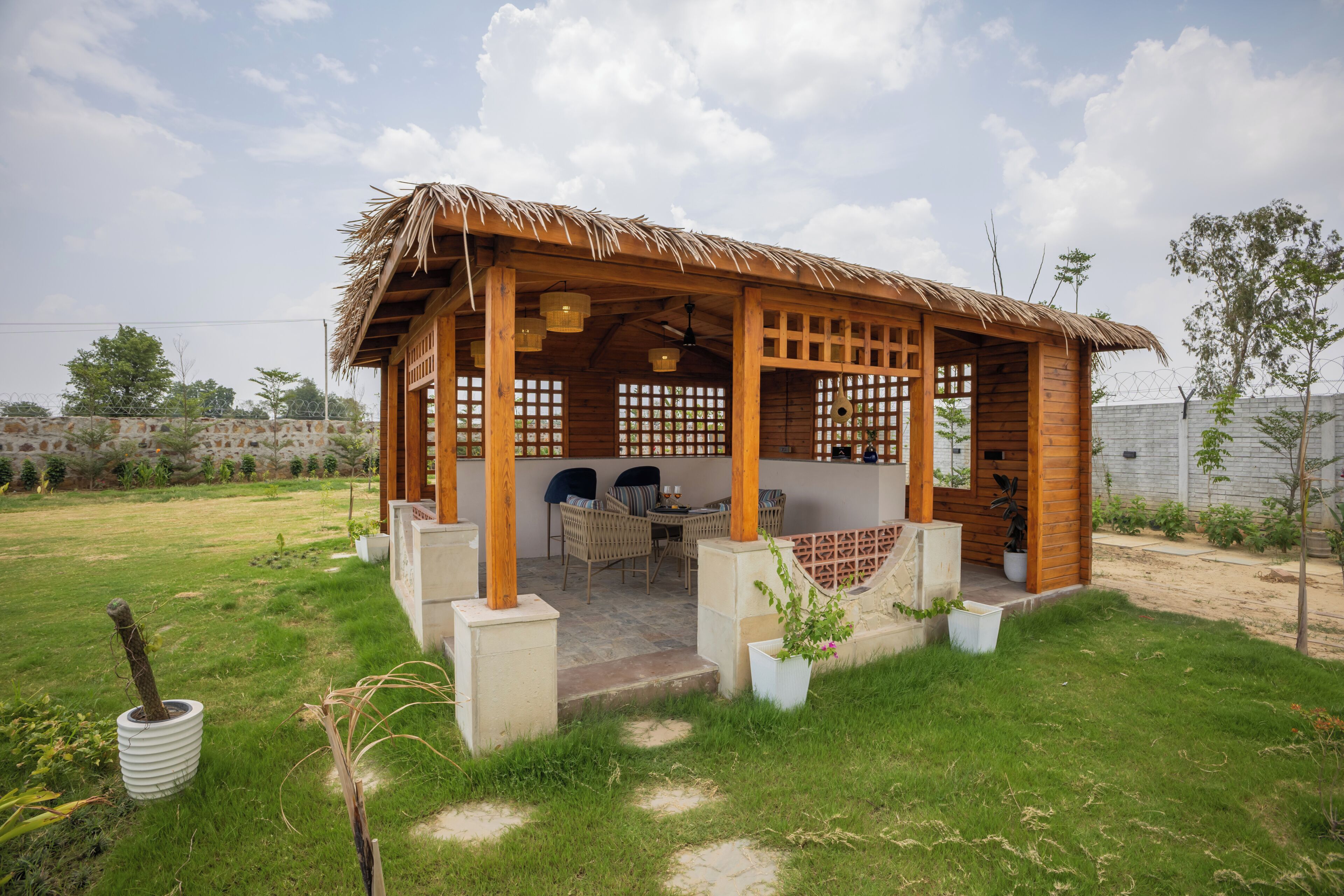 A wooden structure with a thatched roof, a covered seating area with chairs, and a small garden.