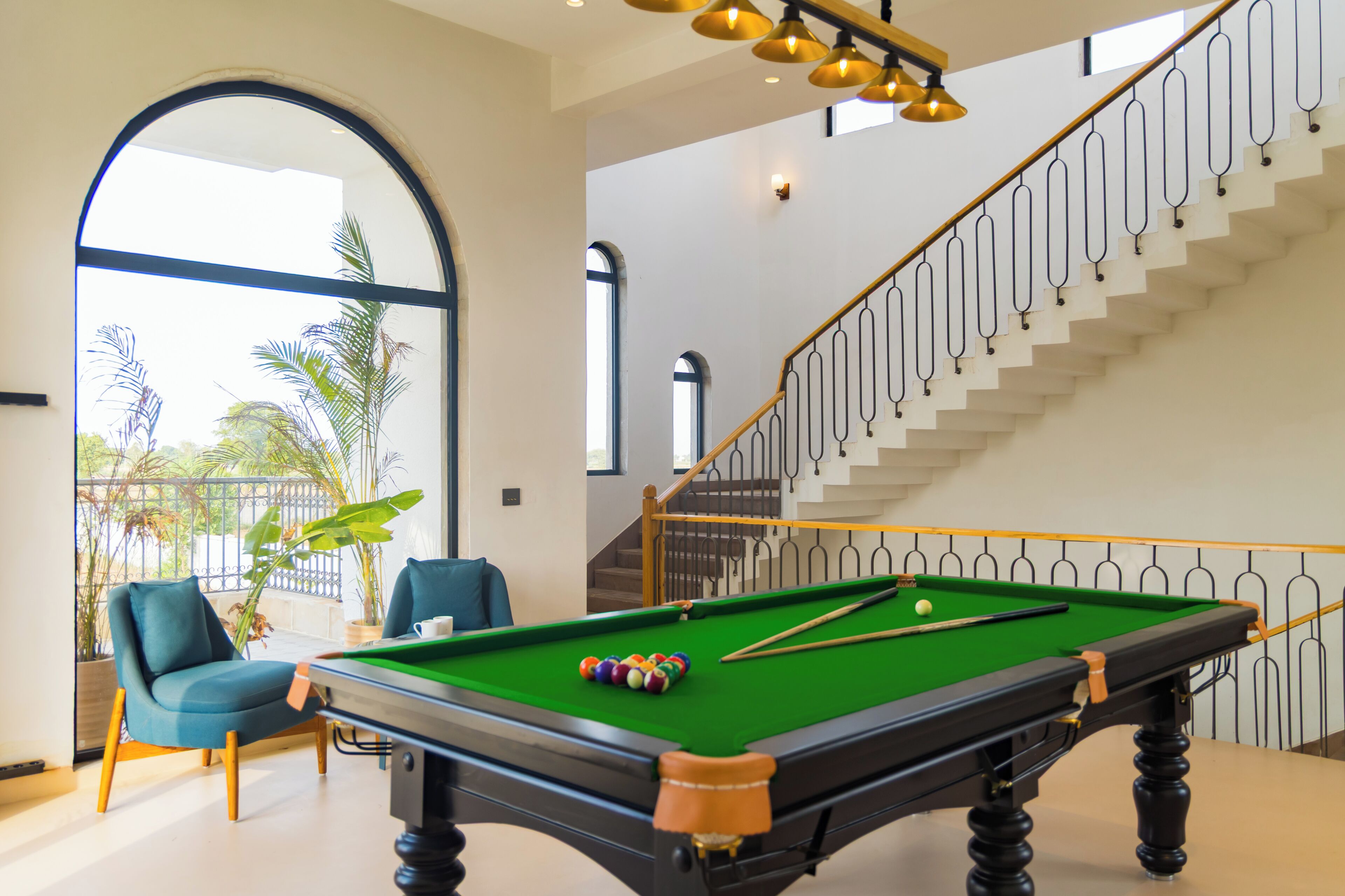 Una mesa de billar en una habitación moderna con una escalera y ventanas amplias.