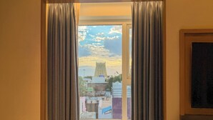 Una habitación con ventana, cortinas, una pequeña mesa con cafetera y vistas a edificios y una torre.