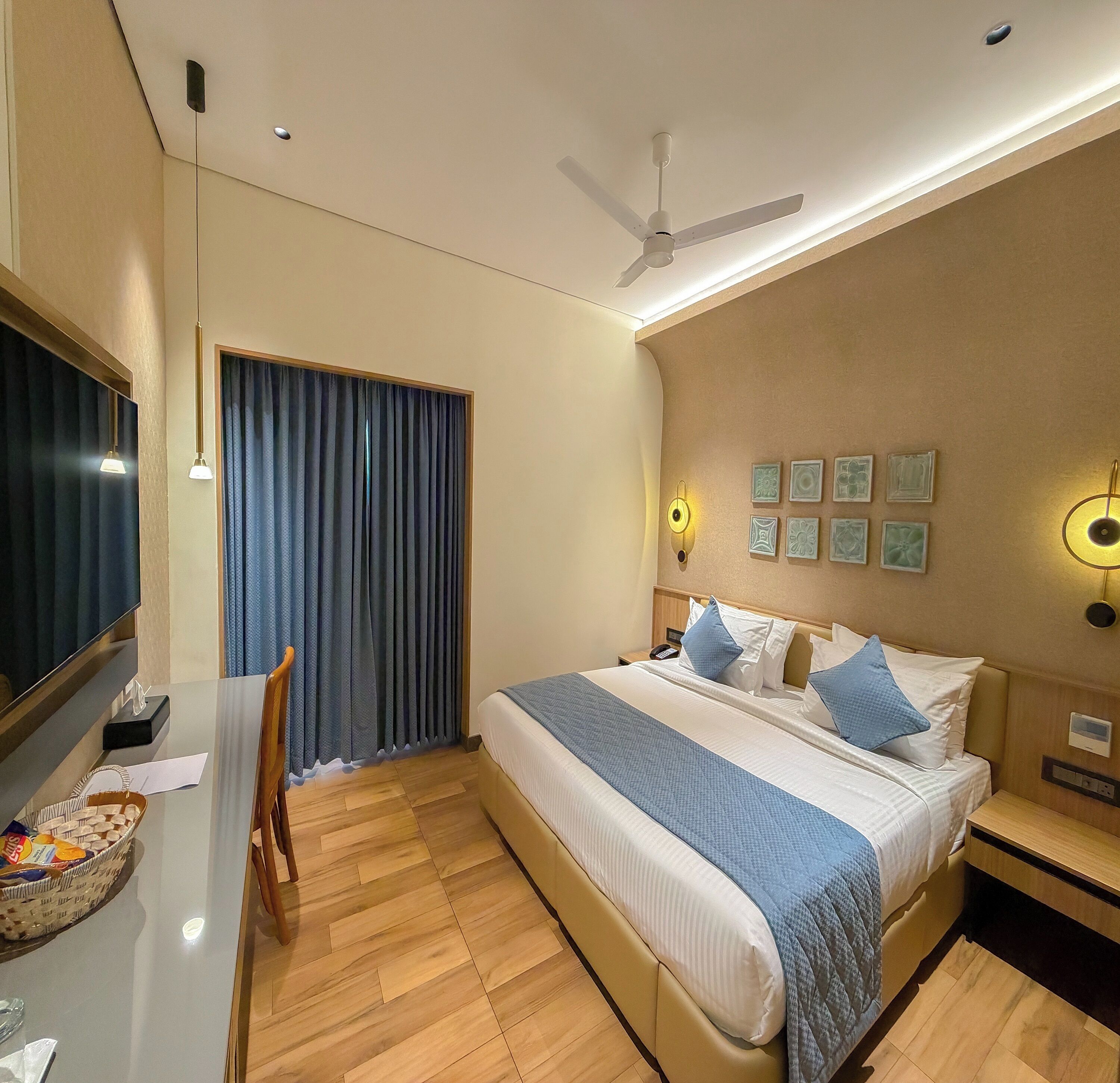 Habitación de hotel con cama, mesita de noche, escritorio, silla, cortinas, ventilador de techo, luz empotrada en la pared y obra de arte.