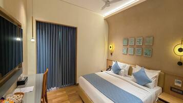 Habitación de hotel con cama, mesita de noche, escritorio, silla, cortinas, ventilador de techo, luz empotrada en la pared y obra de arte.