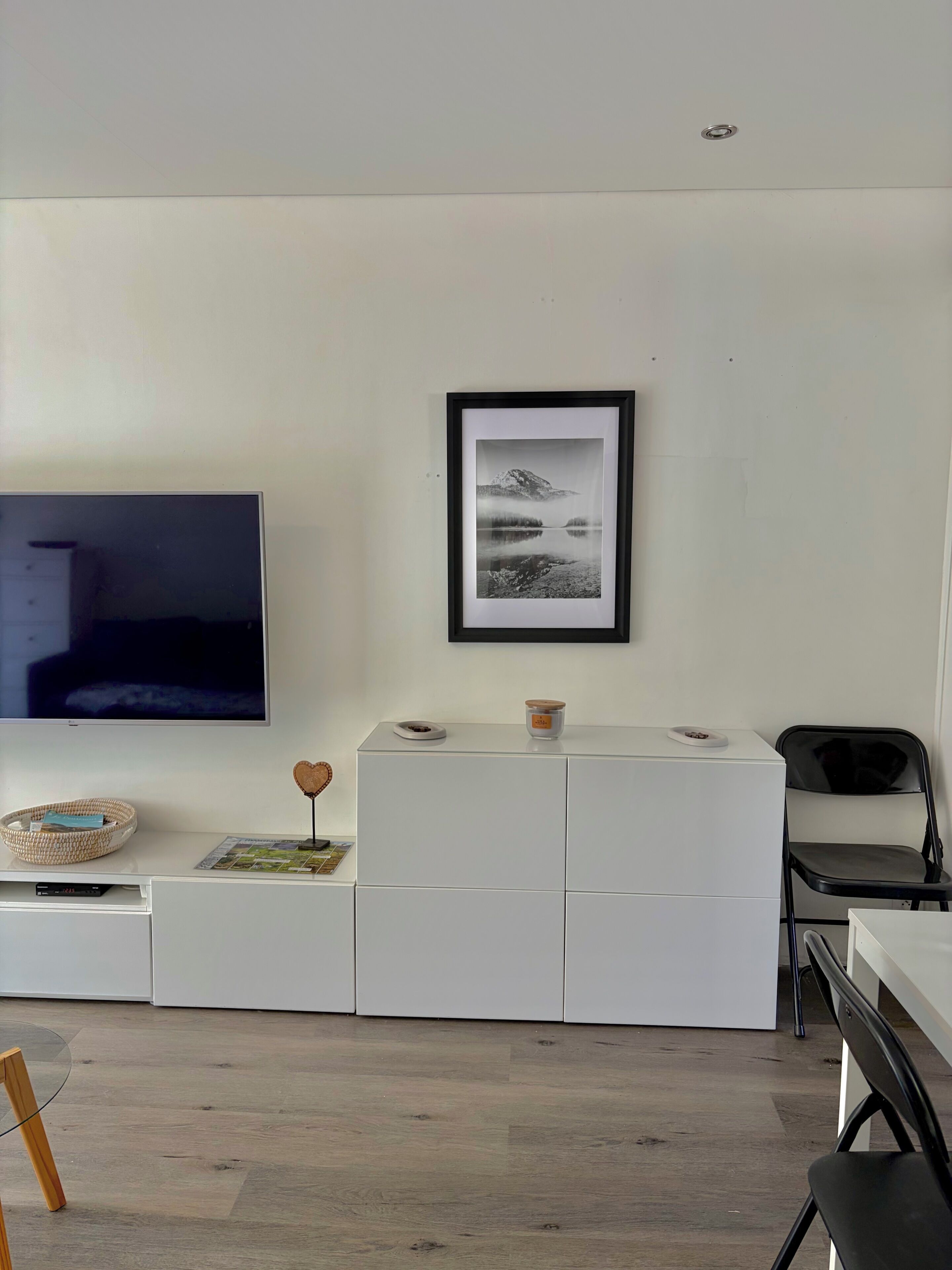 Un soggiorno moderno con una televisione a schermo piatto, una fotografia incorniciata appesa al muro, un mobiletto bianco con un tavolino e una sedia pieghevole nera.