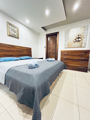 Ein modernes Schlafzimmer mit einem hölzernen Kopfteil, einem großen Bett mit grauer, strukturierter Bettdecke, zwei aufgerollten Handtüchern, eine Holzkommode und eine Tür mit Holzrahmen.