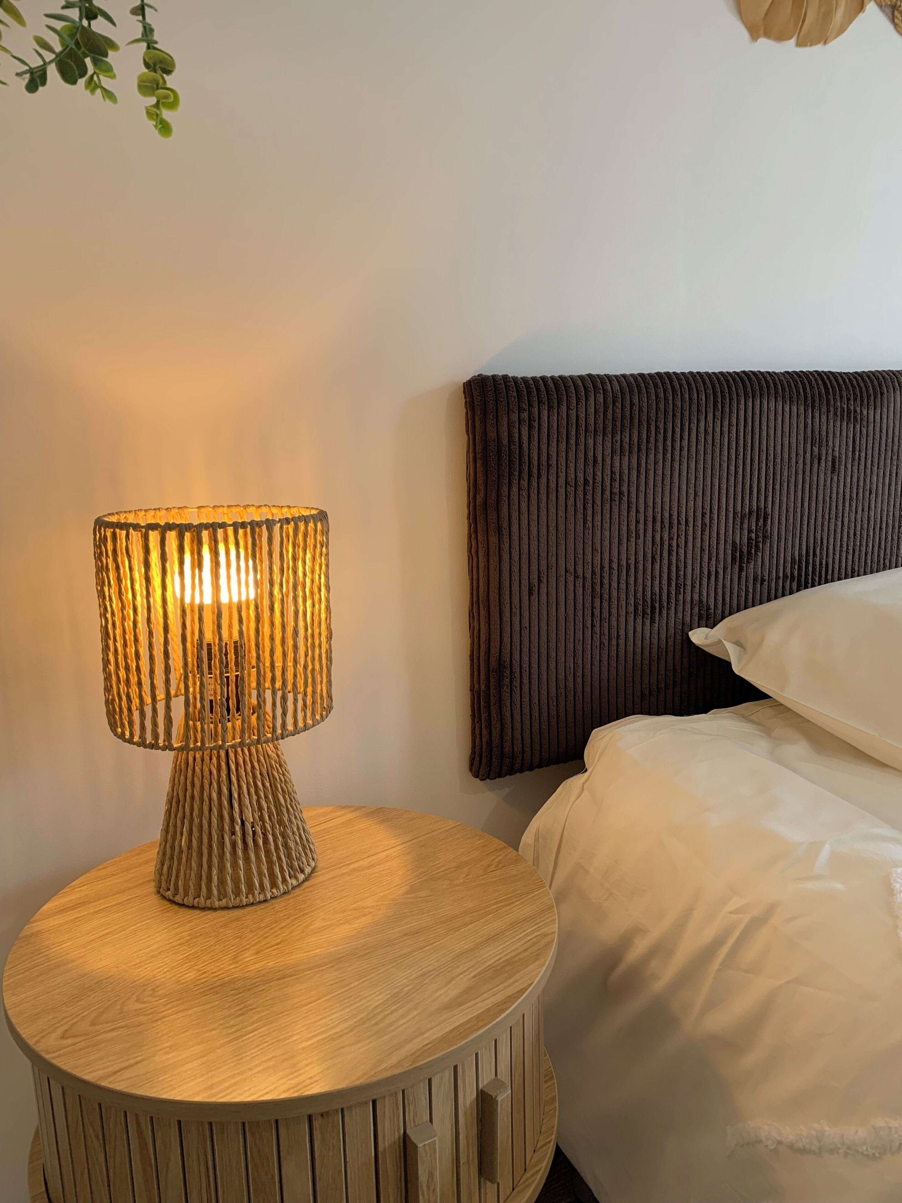 A round wooden nightstand with a textured lamp, a dark headboard, and a bed with white bedding.