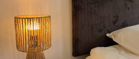 A round wooden nightstand with a textured lamp, a dark headboard, and a bed with white bedding.