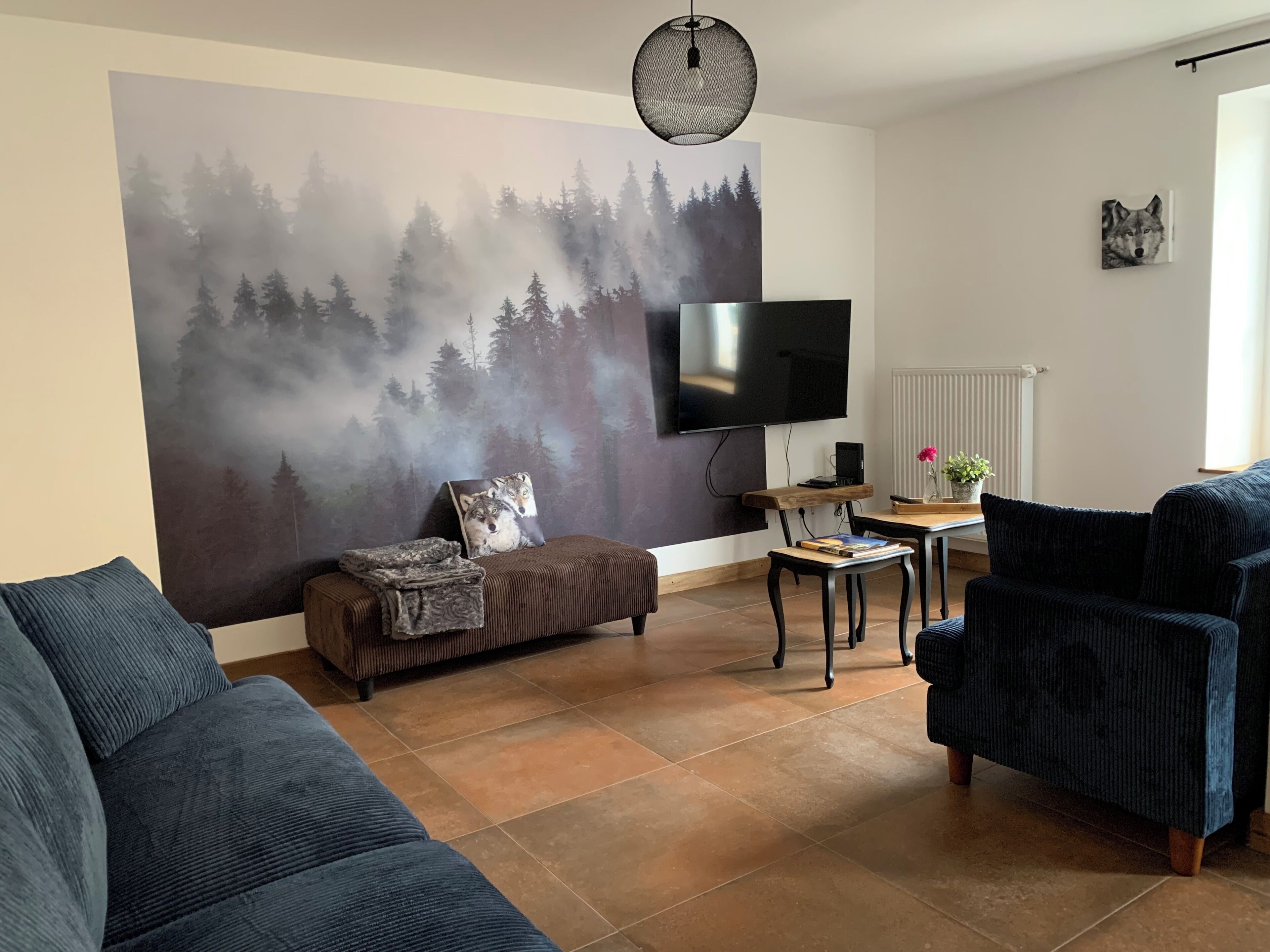 Un salon avec une grande fresque murale, un téléviseur à écran plat, un canapé, un fauteuil et une petite table.