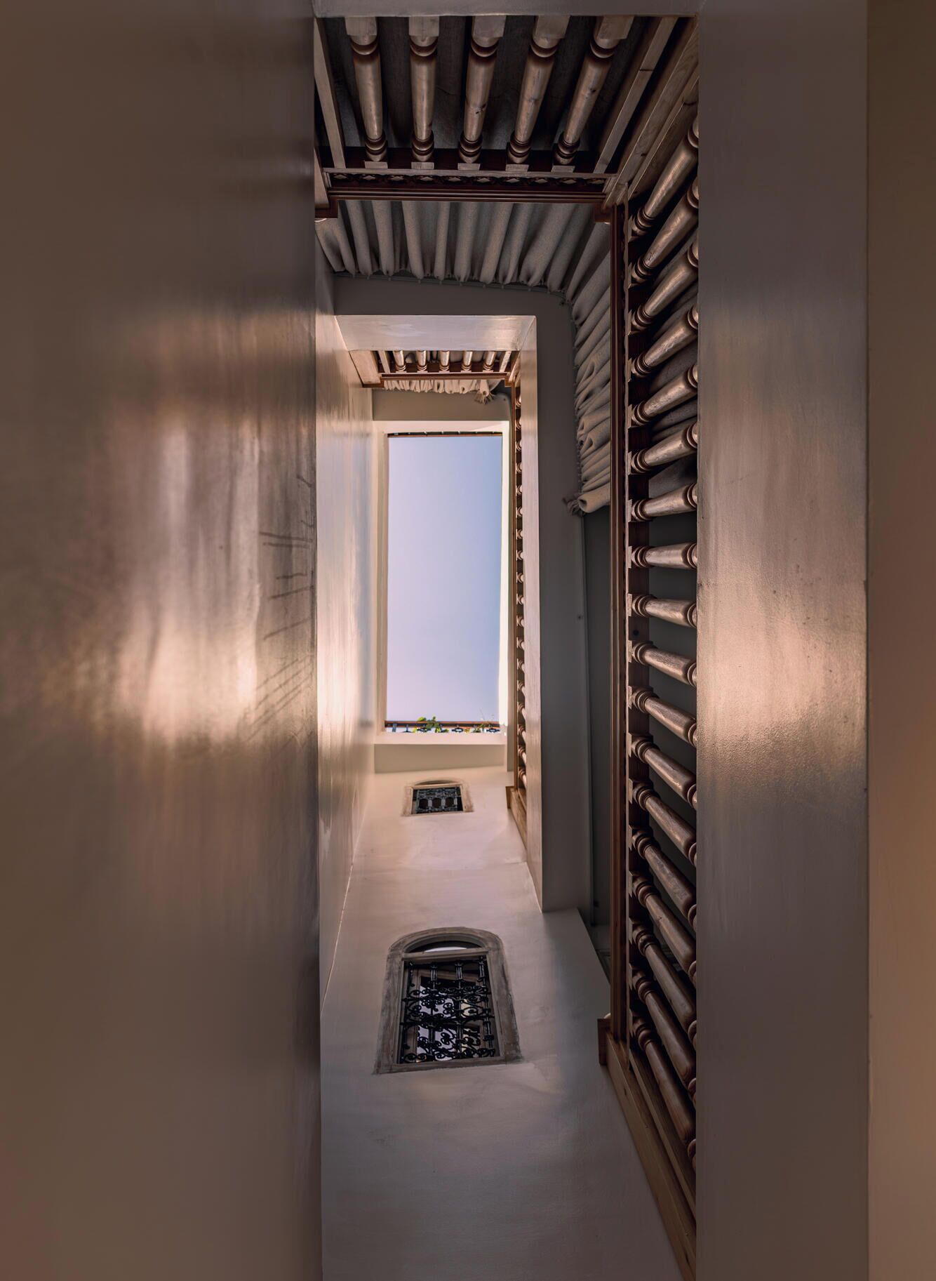 Un couloir étroit, avec une fenêtre au fond, une grille métallique sur le côté droit et une petite grille d’aération dans le sol.