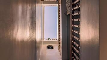 Un couloir étroit, avec une fenêtre au fond, une grille métallique sur le côté droit et une petite grille d’aération dans le sol.