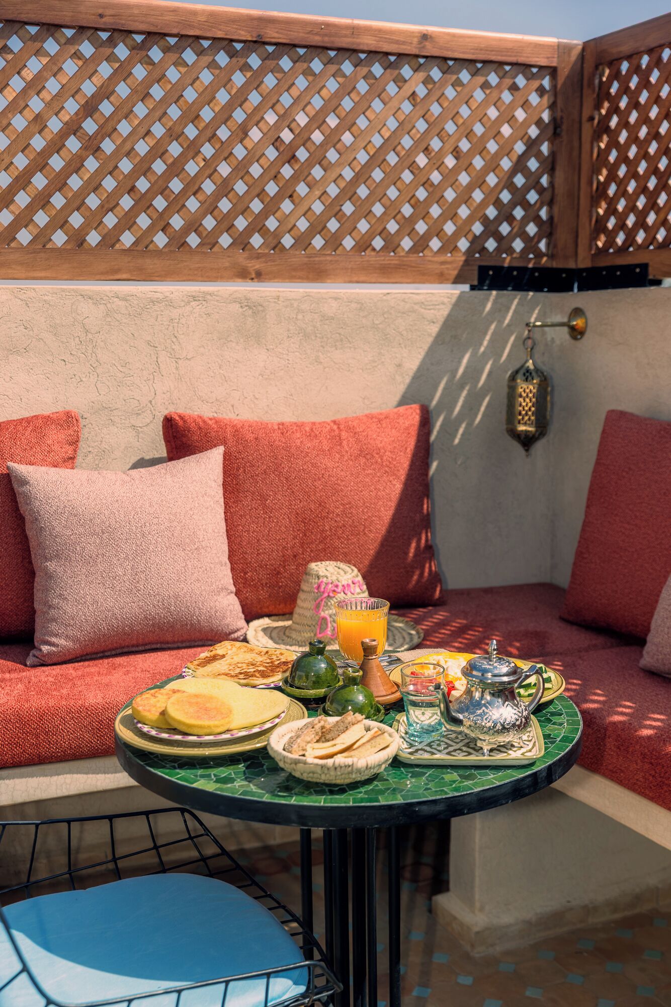 Un espace extérieur accueillant avec des sièges confortables et une table dressée pour un repas, agrémenté d’un panier en osier, d’un verre de jus d’orange et d’un pichet en métal.
