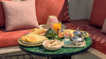 Un espace extérieur accueillant avec des sièges confortables et une table dressée pour un repas, agrémenté d’un panier en osier, d’un verre de jus d’orange et d’un pichet en métal.