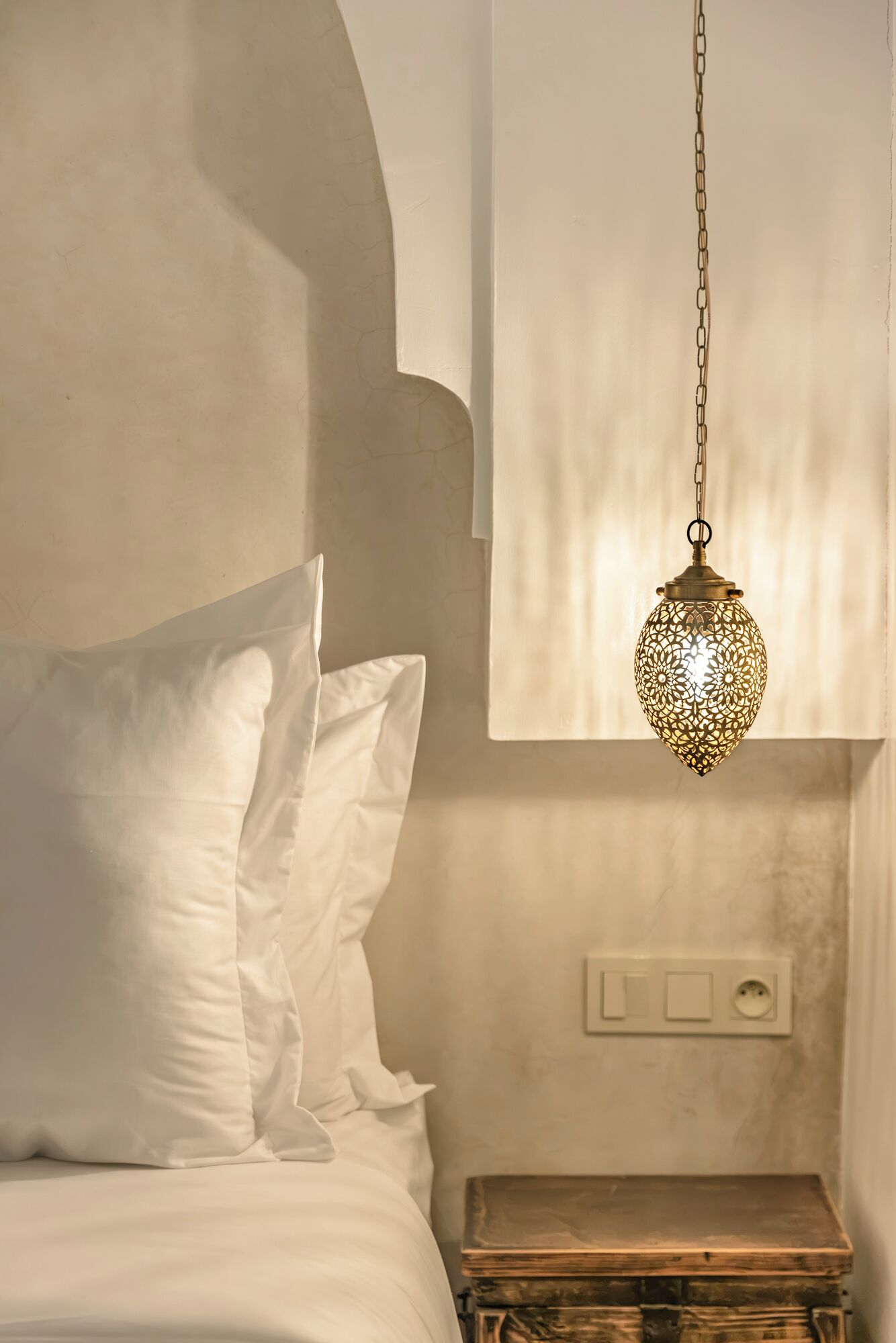 A bedroom with a bed, pillows, a hanging light fixture, and a bedside table.