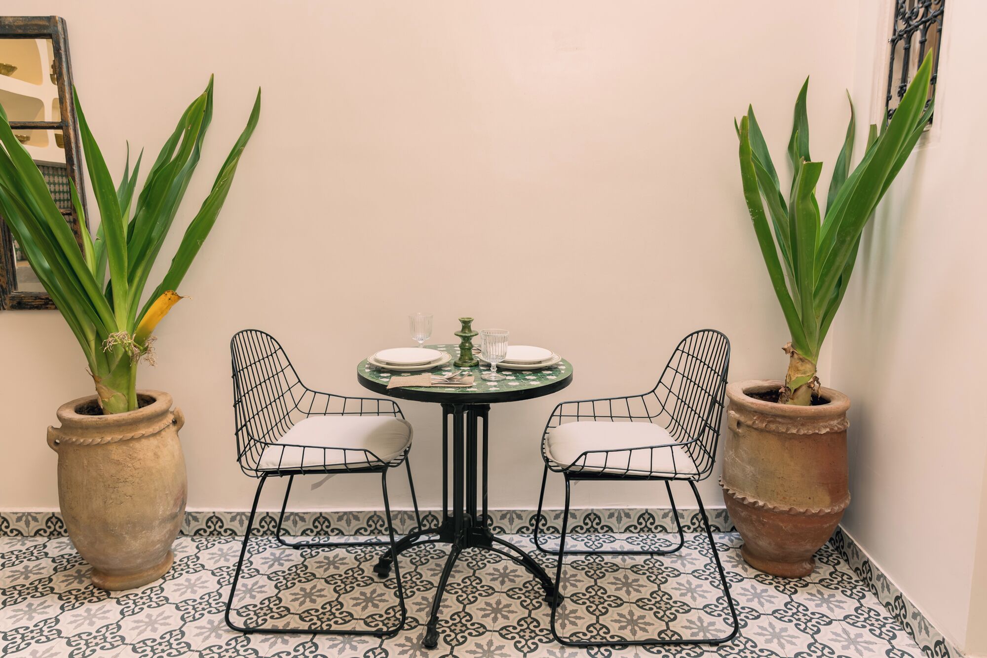 A corner with a round table set for two, two chairs, two potted plants, and a patterned floor.