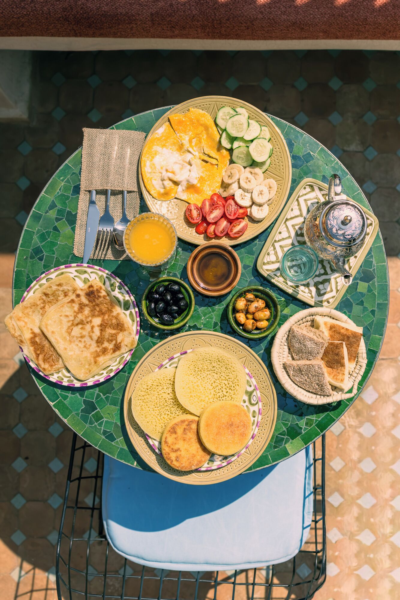 A table with a variety of breakfast items including flatbreads, a plate with sliced tomatoes, cucumbers, and bananas, a bowl of olives, and a teapot.