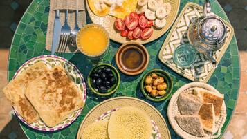 A table with a variety of breakfast items including flatbreads, a plate with sliced tomatoes, cucumbers, and bananas, a bowl of olives, and a teapot.
