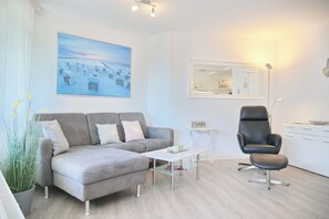 Ein modernes Wohnzimmer mit einem grauen Sofa, einem schwarzen Sessel, einem kleinen weißen Tisch und einem großen Gemälde, das eine Strandansicht zeigt.