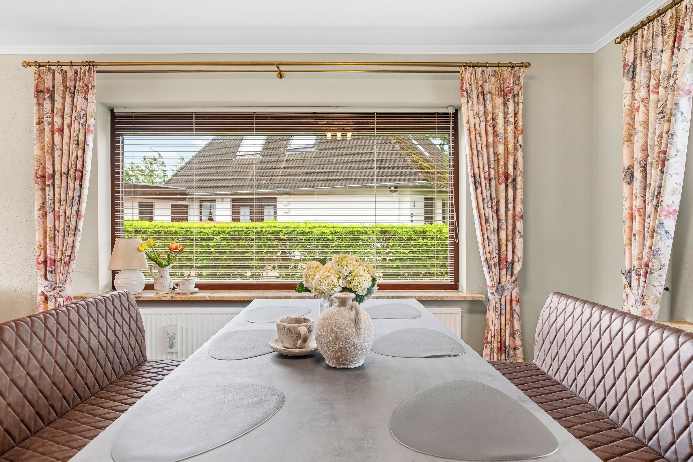 Ein Essbereich mit einem für zwei Personen gedecktem Tisch, flankiert von gepolsterten Bänken, einer Vase mit Blumen und einem Fenster mit Jalousien.