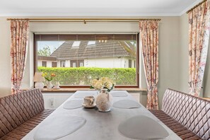 Ein Essbereich mit einem für zwei Personen gedecktem Tisch, flankiert von gepolsterten Bänken, einer Vase mit Blumen und einem Fenster mit Jalousien.